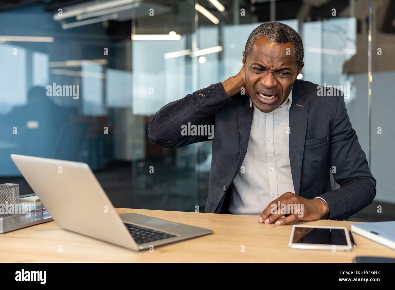 Mature man working on a laptop experiencing discomfort and strain in his neck, holding it with his hand while feeling pain from poor posture or prolonged computer use in an office setting Stock Photo