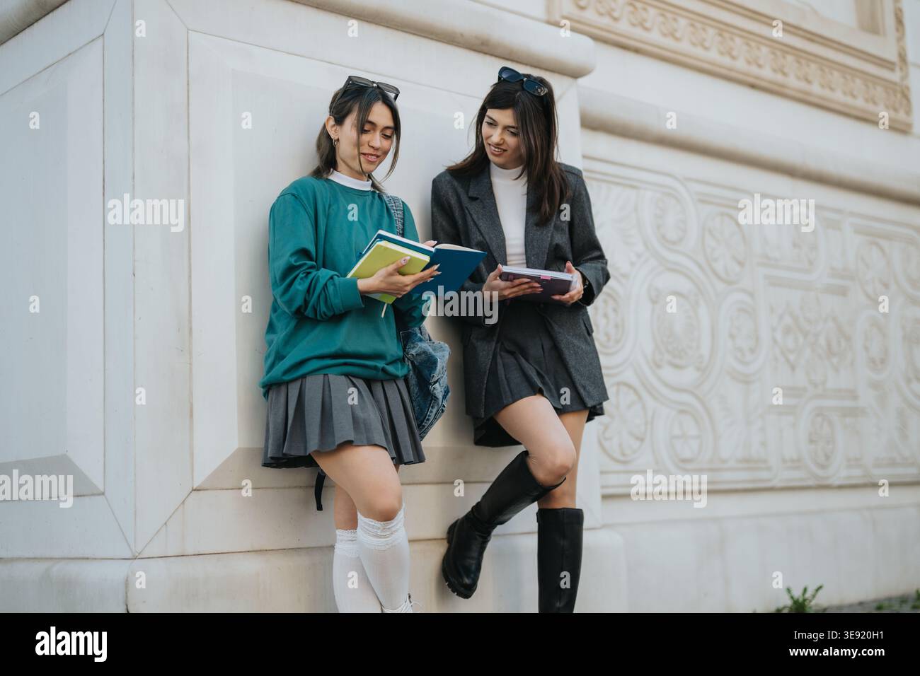 Two female students studying and chatting outdoors by decorative building wall with books and backpacks Stock Photo