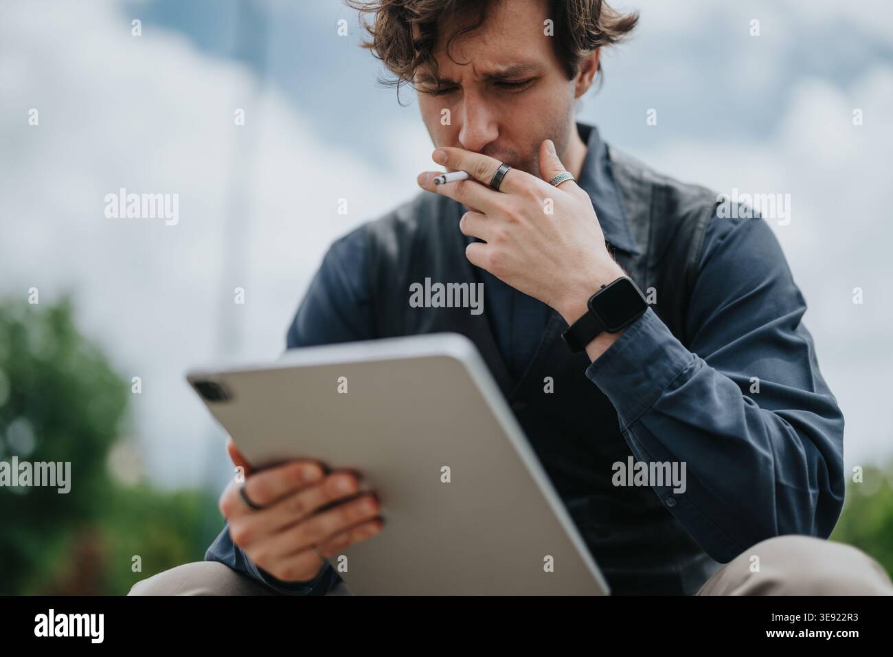 Businessman smoking while reading tablet outdoors, thoughtful young professional using digital device Stock Photo