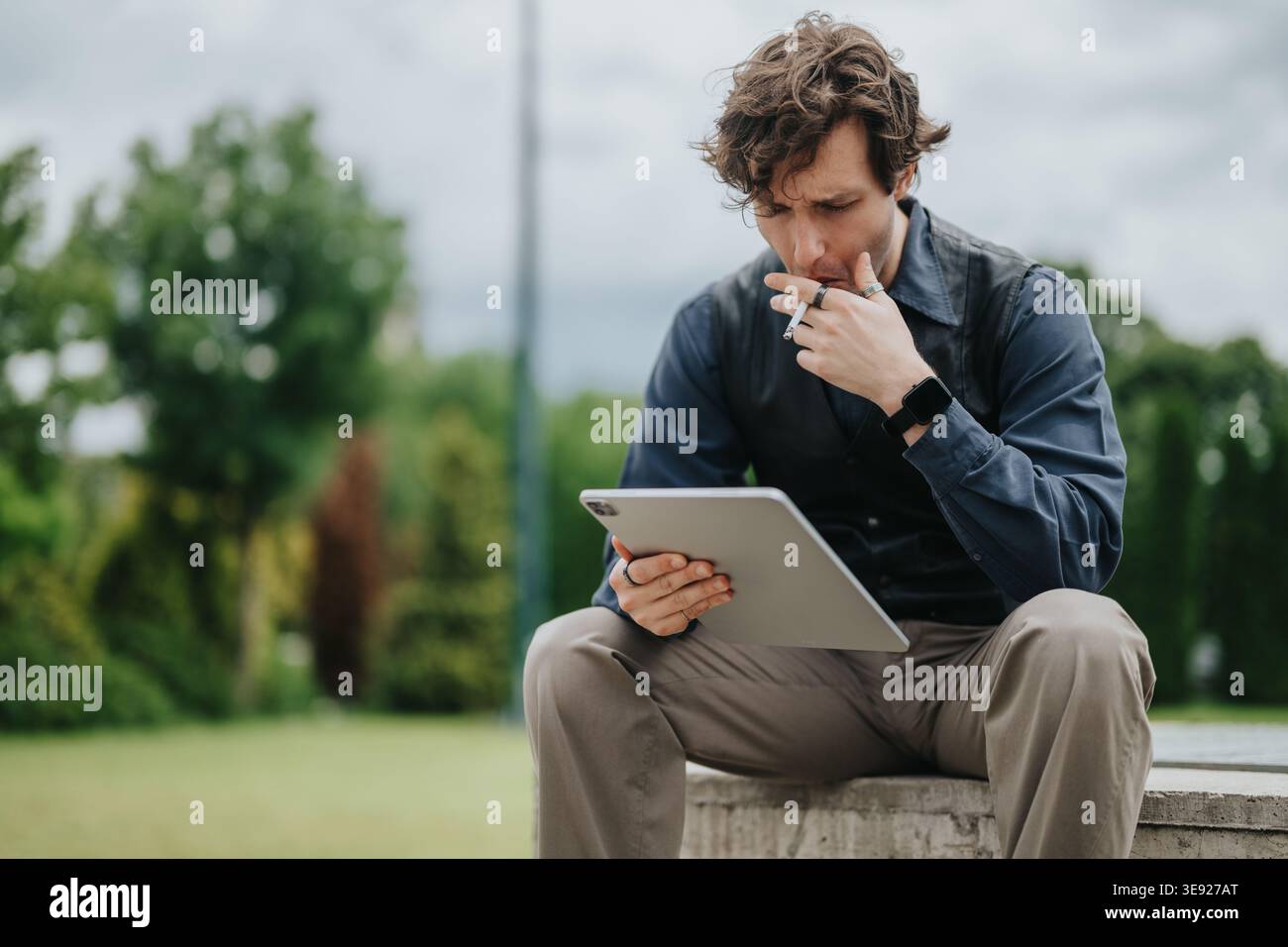 Businessman smoking while using a tablet outdoors, looking thoughtful during a park break Stock Photo