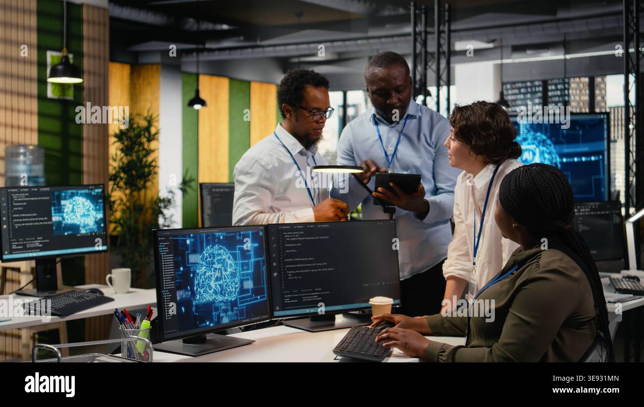 Multiethnic technicians use devices in AI startup office to train predictive models for language processing tasks. Multiracial coworkers use deep learning technical expertise for tasks, camera A Stock Photo