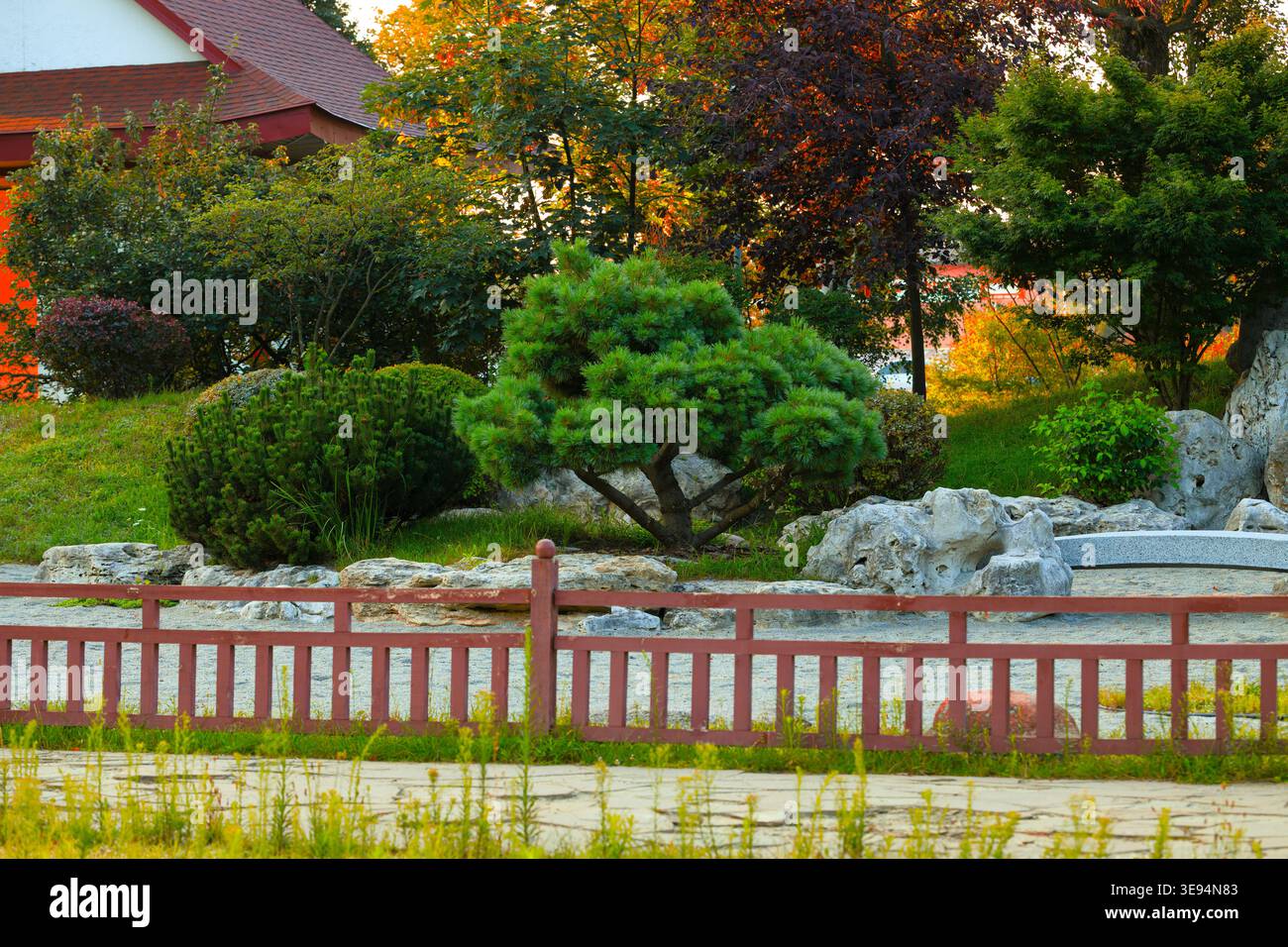 A tranquil Japanese garden scene featuring a red wooden fence, lush greenery, and vibrant autumn-colored trees, with large rocks and a stone path Stock Photo