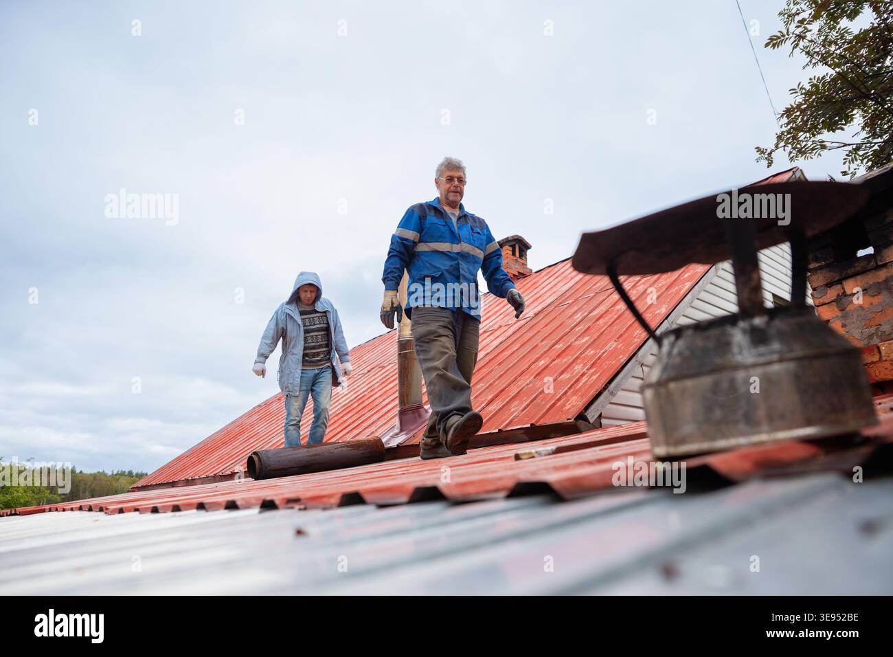 Two men repairs chimney on house roof. Roofer working on metal rooftop construction for home maintenance. Professional worker repairing ventilation sy Stock Photo