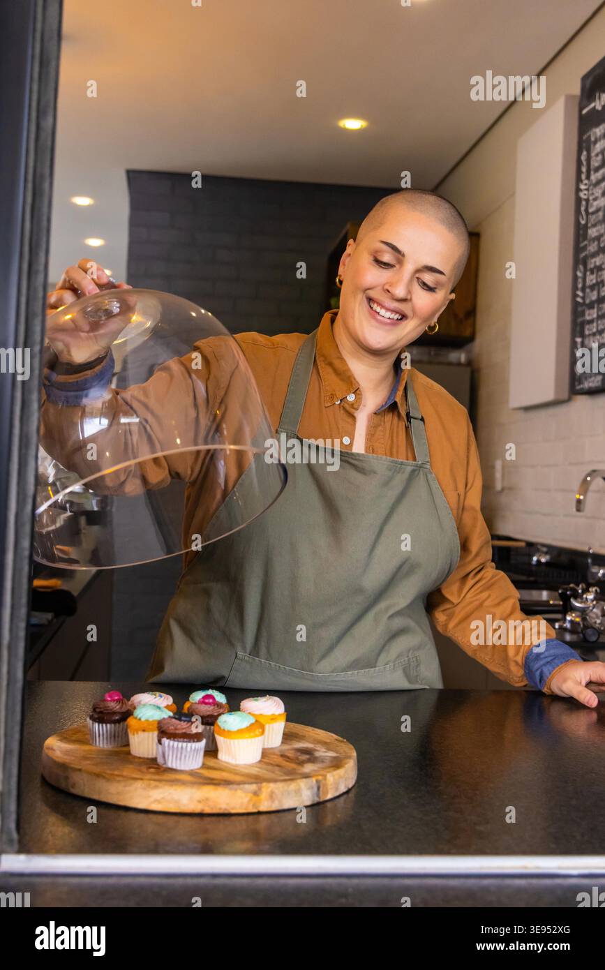 Female baker in apron lifting glass cloche, revealing frosted cupcakes on board at bakery counter. Pastries, dessert, cafe, warm, colorful, chalkboard, espresso Stock Photo