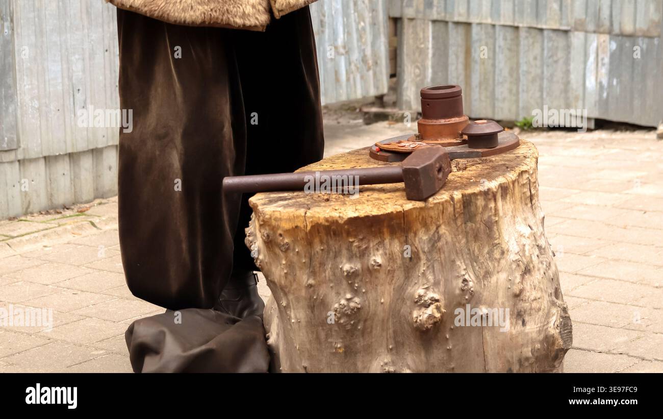 Close-up of a large old hammer lying on a tree stump anvil. Historical reenactment of traditional coin minting or blacksmithing outdoors. Craftsmans t Stock Photo