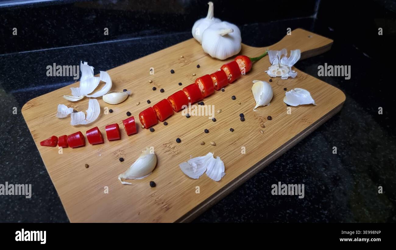 A sliced red chili pepper lies next to peeled garlic cloves and pepper corns on a wooden chopping board. - Smartphone Captured Stock Image