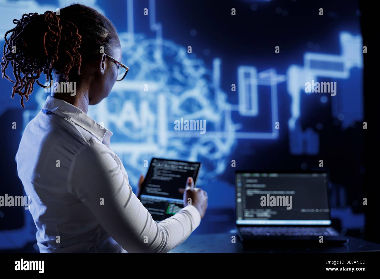 BIPOC engineer updating neural networks made up of interconnected nodes, writing intricate binary code scripts on laptop and tablet. Computer operator using programming to upgrade AI simulation model Stock Photo