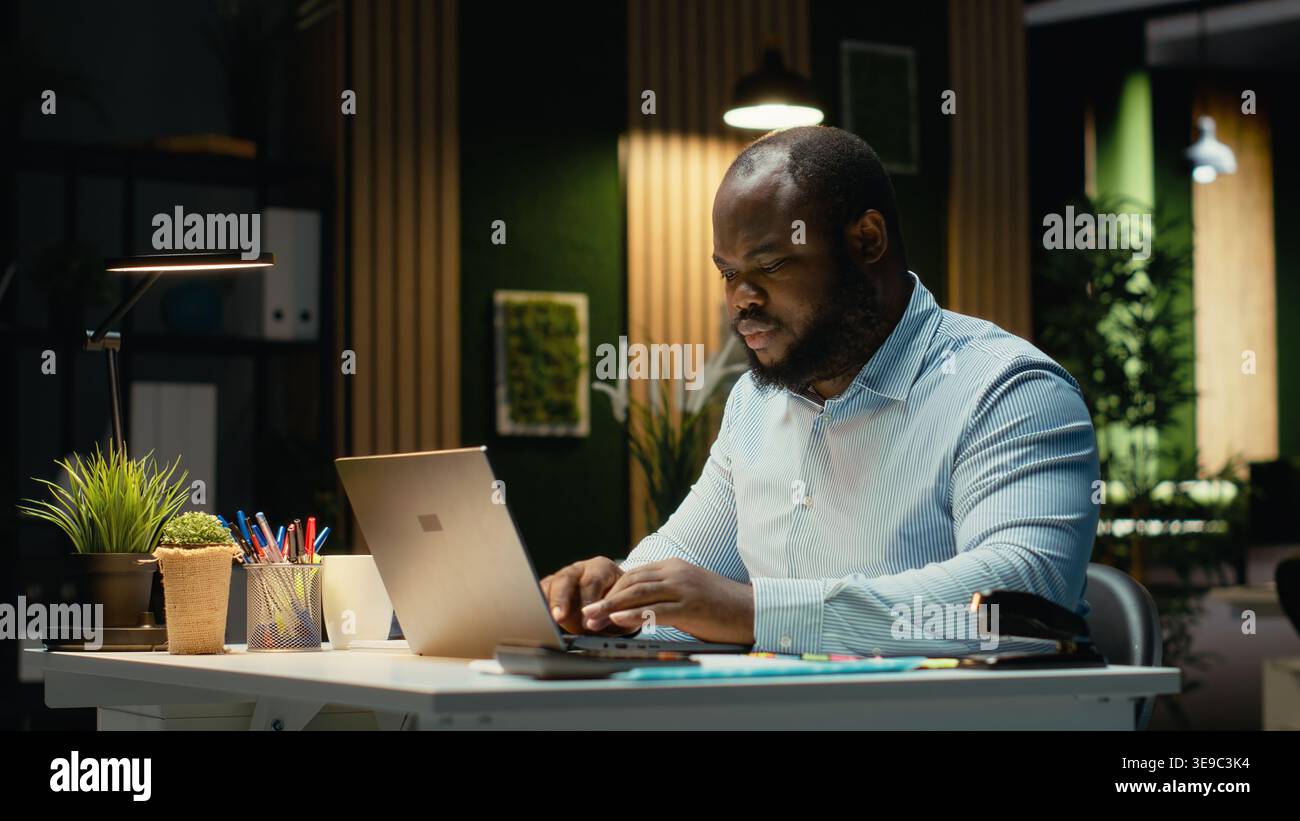 African american guy proofreading a document before transcribing on a textbook, extracting the main ideas from reports on laptop. Expert documenting after hours late at night. Stock Photo