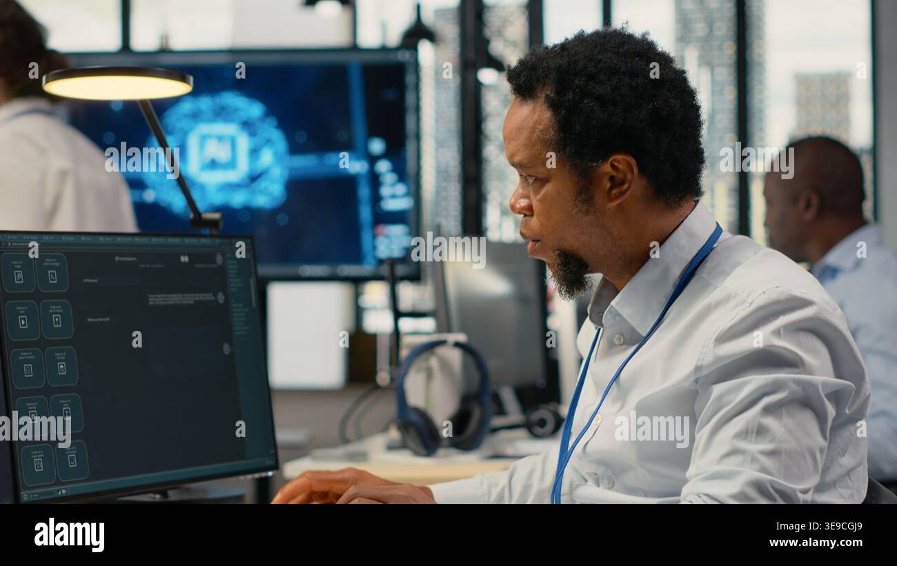 Technician in office solving tasks with artificial intelligence LLM chatbot answering prompts. African american man at work using PC to interact with large language model program, camera B Stock Photo