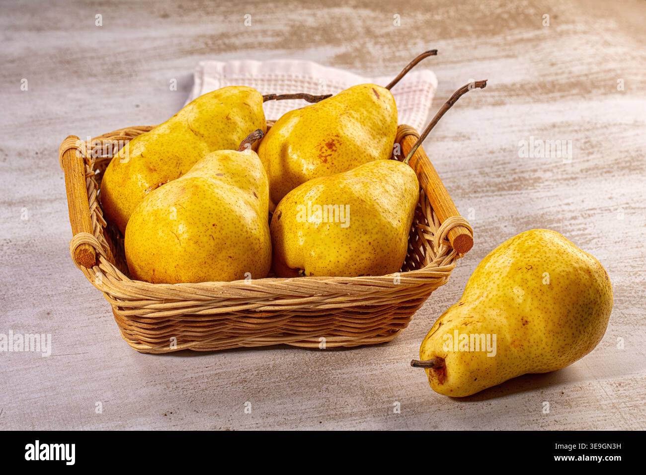 Vibrant yellow pears arranged in rustic wicker basket, one pear rests beside it on light textured wooden surface. Soft natural light creates warm, inv Stock Photo