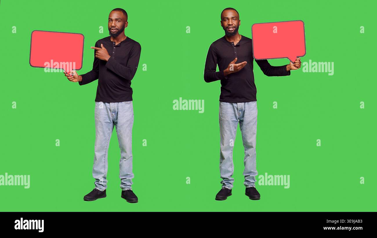Male model holding red blank speech bubble on camera, creating presentation with empty copyspace cardboard icon. Young adult showing mockup billboard over green screen backdrop. Stock Photo