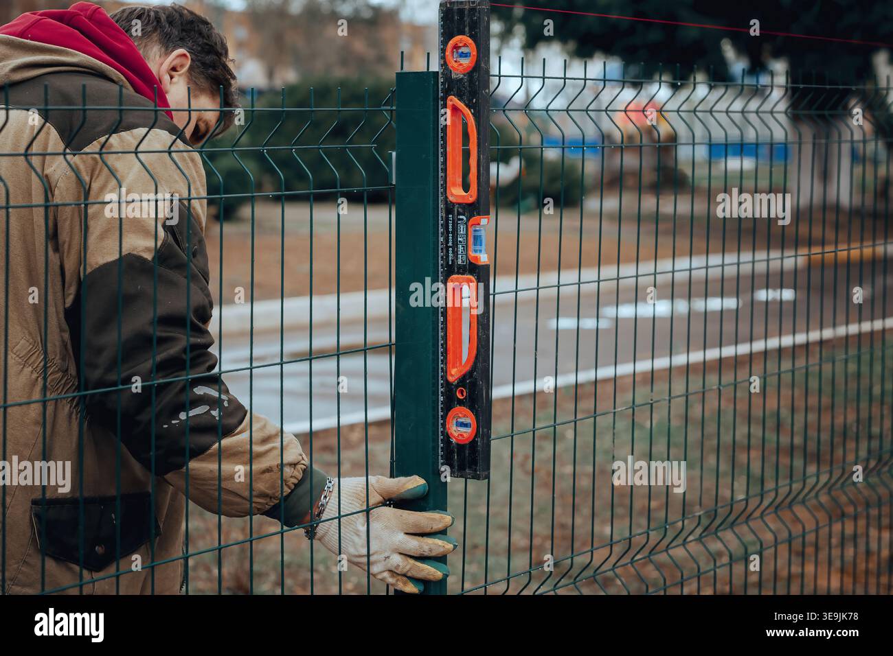 Worker checking vertical alignment of metal fence post using level tool during installation process. Stock Photo