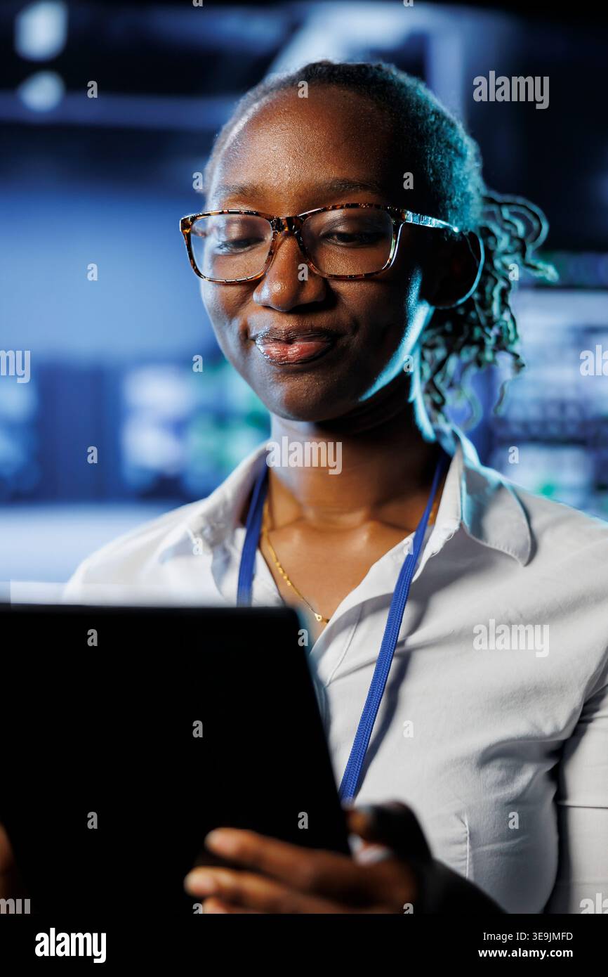 Satisfied admin in data center uses tablet to prevent system electronics overload. Efficient expert between server mainframes ensuring enough network bandwidth for smooth operations Stock Photo