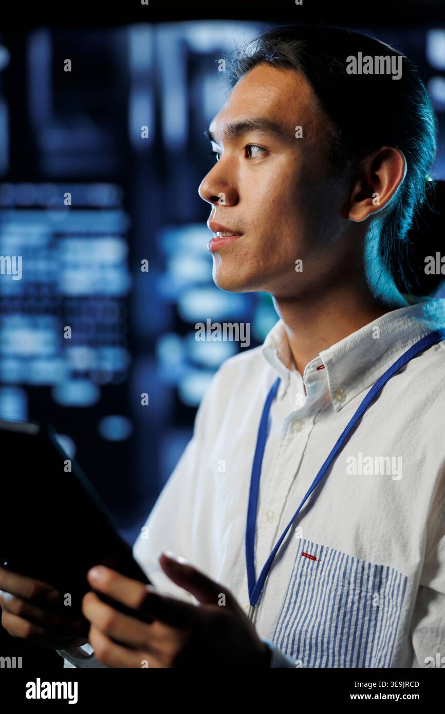 Developer in high tech facility uses tablet to prevent system overload during peak traffic periods. Asian programmer between server rows ensuring enough network bandwidth for smooth operations Stock Photo