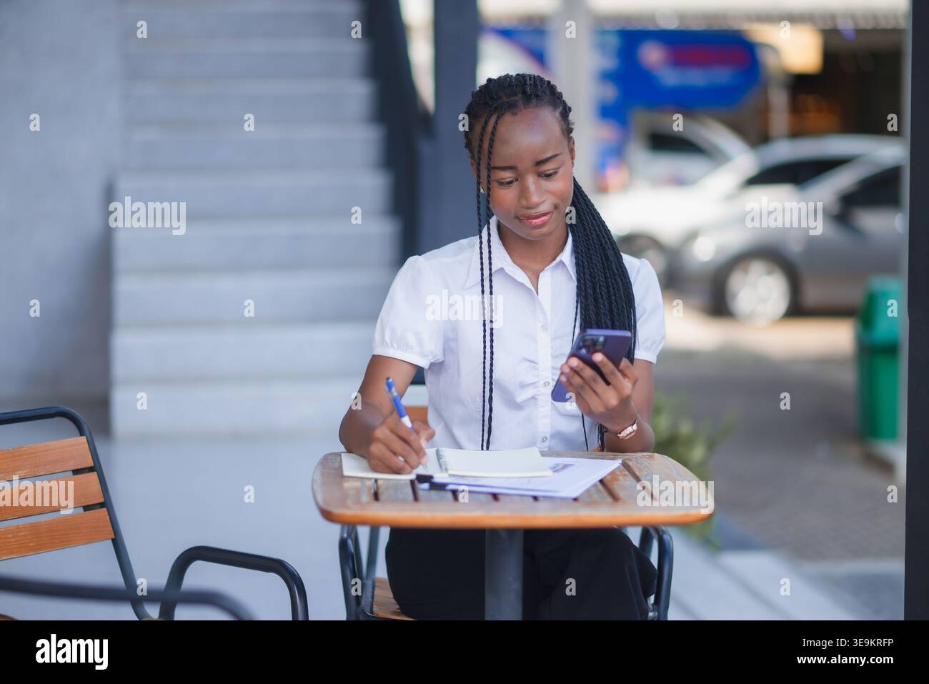 Young Black business woman writing notes and checking smartphone at an outdoor cafe table. Remote work, mobile communication, task planning, productiv Stock Photo