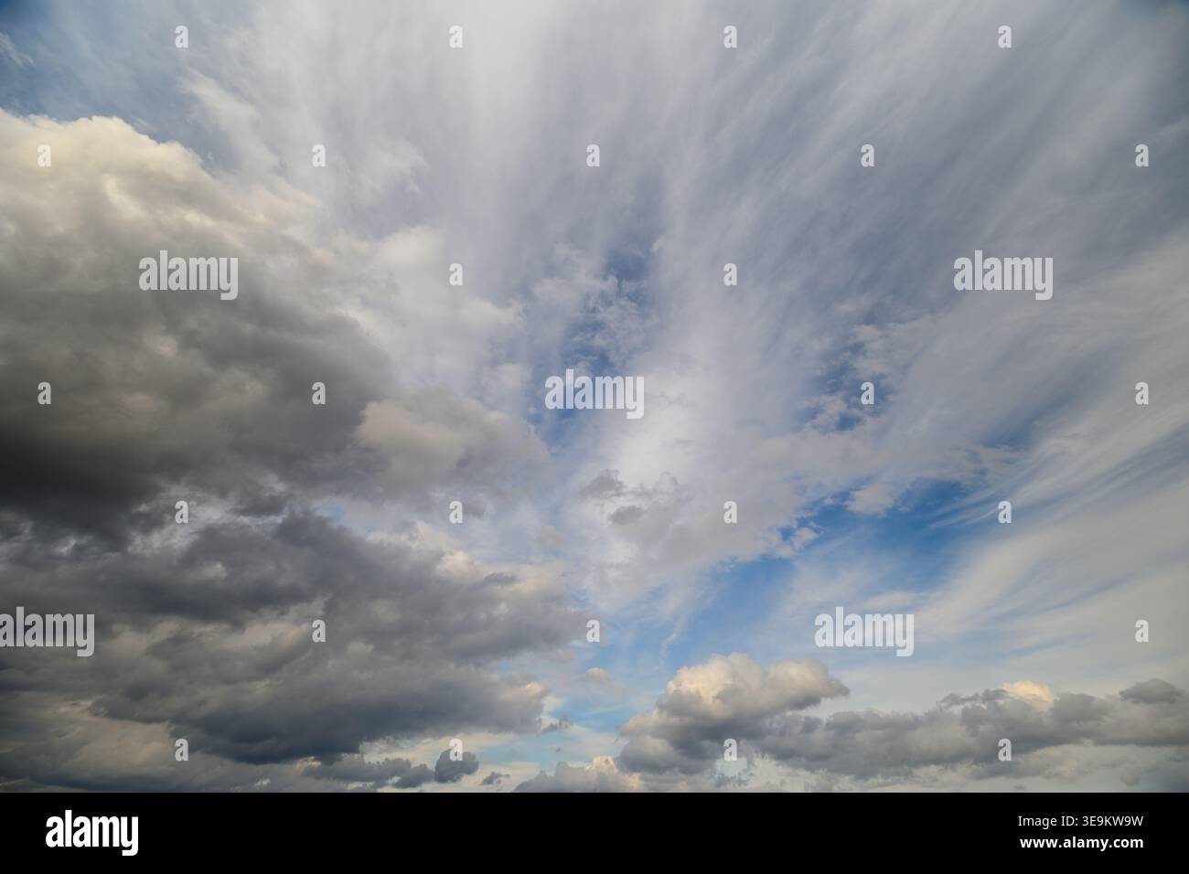 Dramatic layered clouds sweeping across sky with textured cirrus and heavy cumulus, sunrays piercing through breaks, looming stormfront over distant h Stock Photo