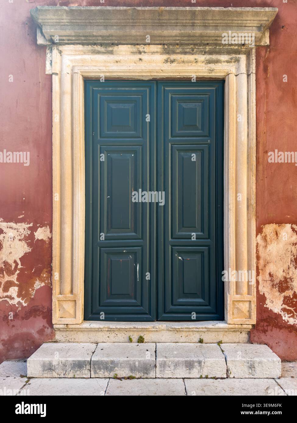 Green wooden double door framed by ornate stonework on a weathered red plaster wall, in Corfu, Ionian Islands, Greece. Stock Photo