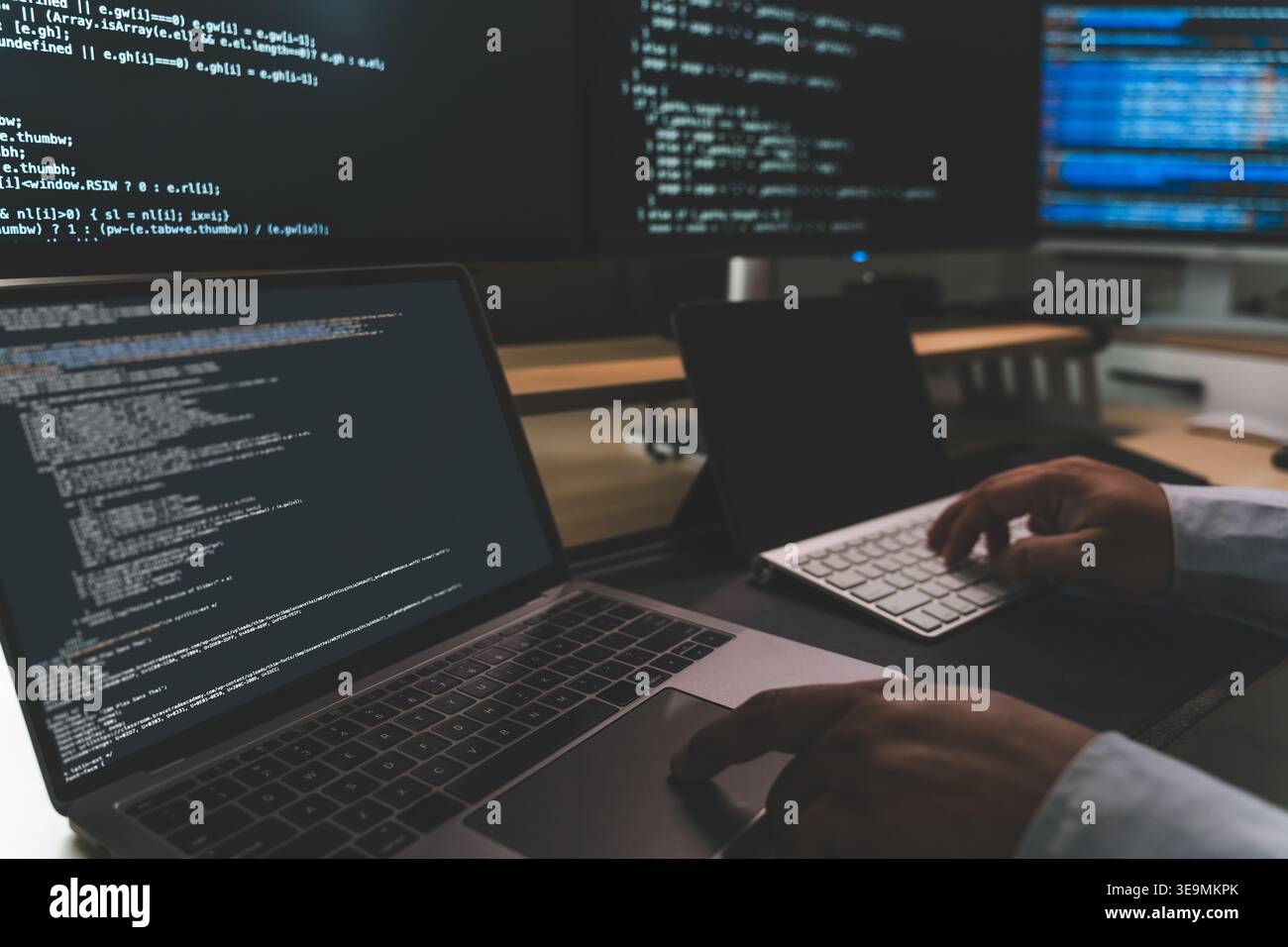 Close up of coding workstation with laptop and multiple screens displaying software code, showing programming workflow, software development, technica Stock Photo
