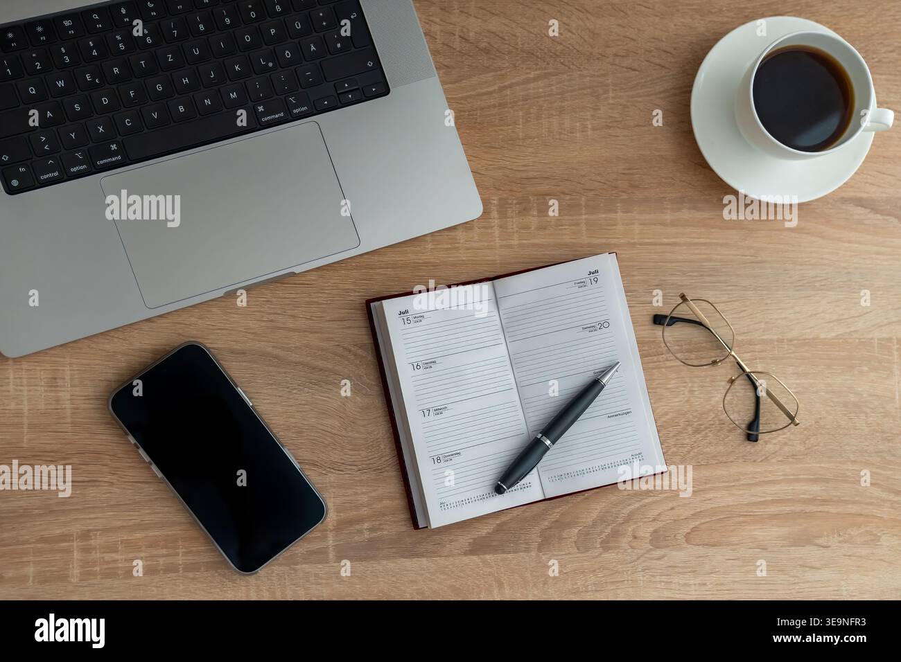 Small planner beside coffee and laptop, pen resting on open weekly spread, folded glasses and textured wood surface create calm inspired mood for idea Stock Photo