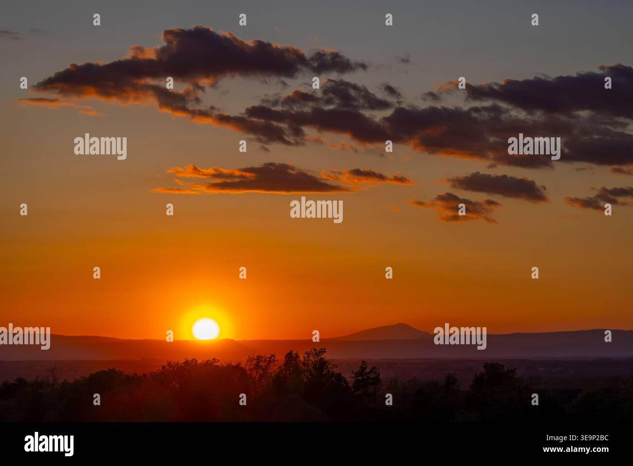 Sun setting behind distant mountains and silhouetted trees with orange sky and clouds Stock Photo