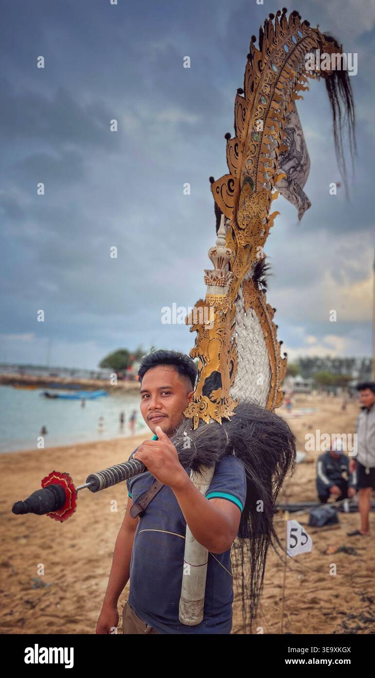 A Balinese man carrying a large traditional wooden windmill propeller decorated with intricate carvings for a local competition on Sanur beach, Bali - Smartphone Captured Stock Image