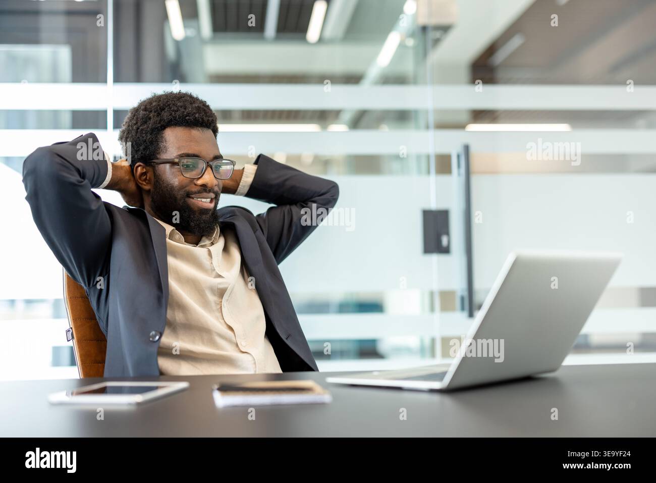 Young professional man with a beard and eyeglasses leaning back in his chair, smiling and looking at a laptop while taking a break from work in a bright, contemporary office environment Stock Photo