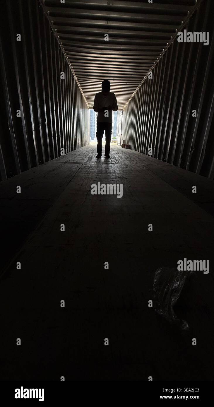A silhouette of a person standing at the far end of a dark industrial shipping container, backlit by bright daylight, creating a dramatic and moody at - Smartphone Captured Stock Image