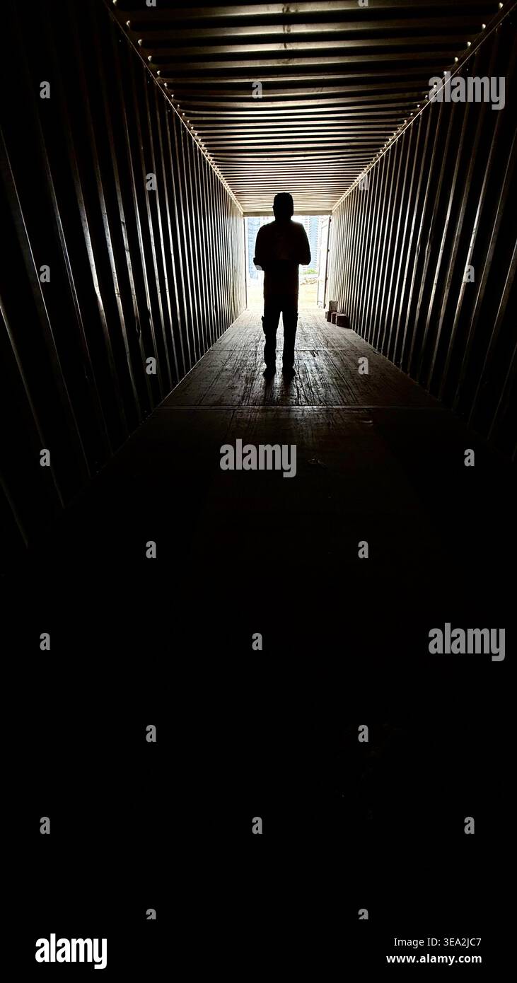A silhouette of a person standing at the far end of a dark industrial shipping container, backlit by bright daylight, creating a dramatic and moody at - Smartphone Captured Stock Image