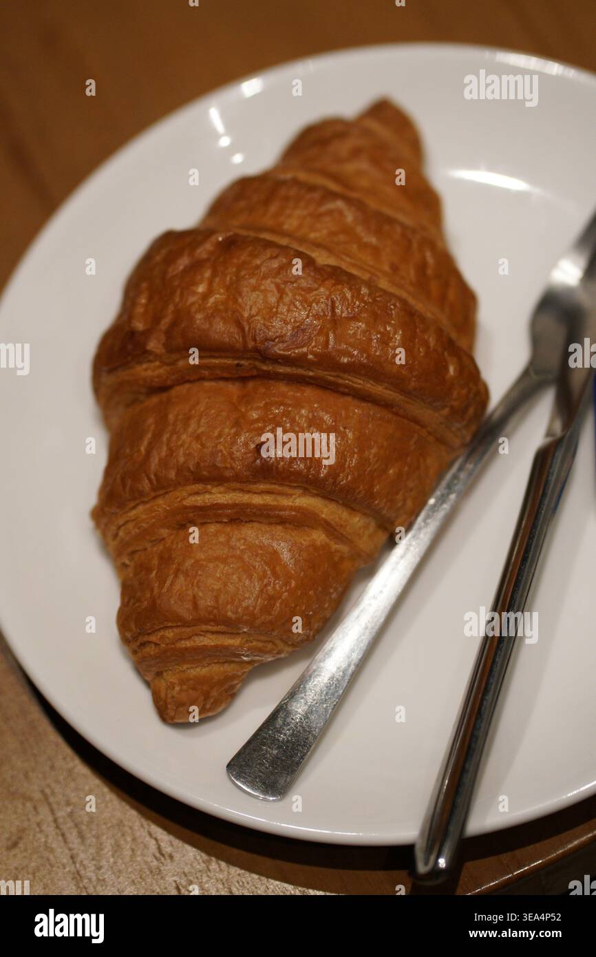 Golden-brown croissant on white plate with knife and fork, placed on wooden surface. Stock Photo