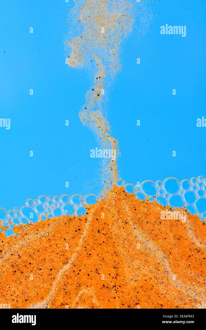 Stream orange sand falling into liquid with bubbles against bright blue background Stock Photo