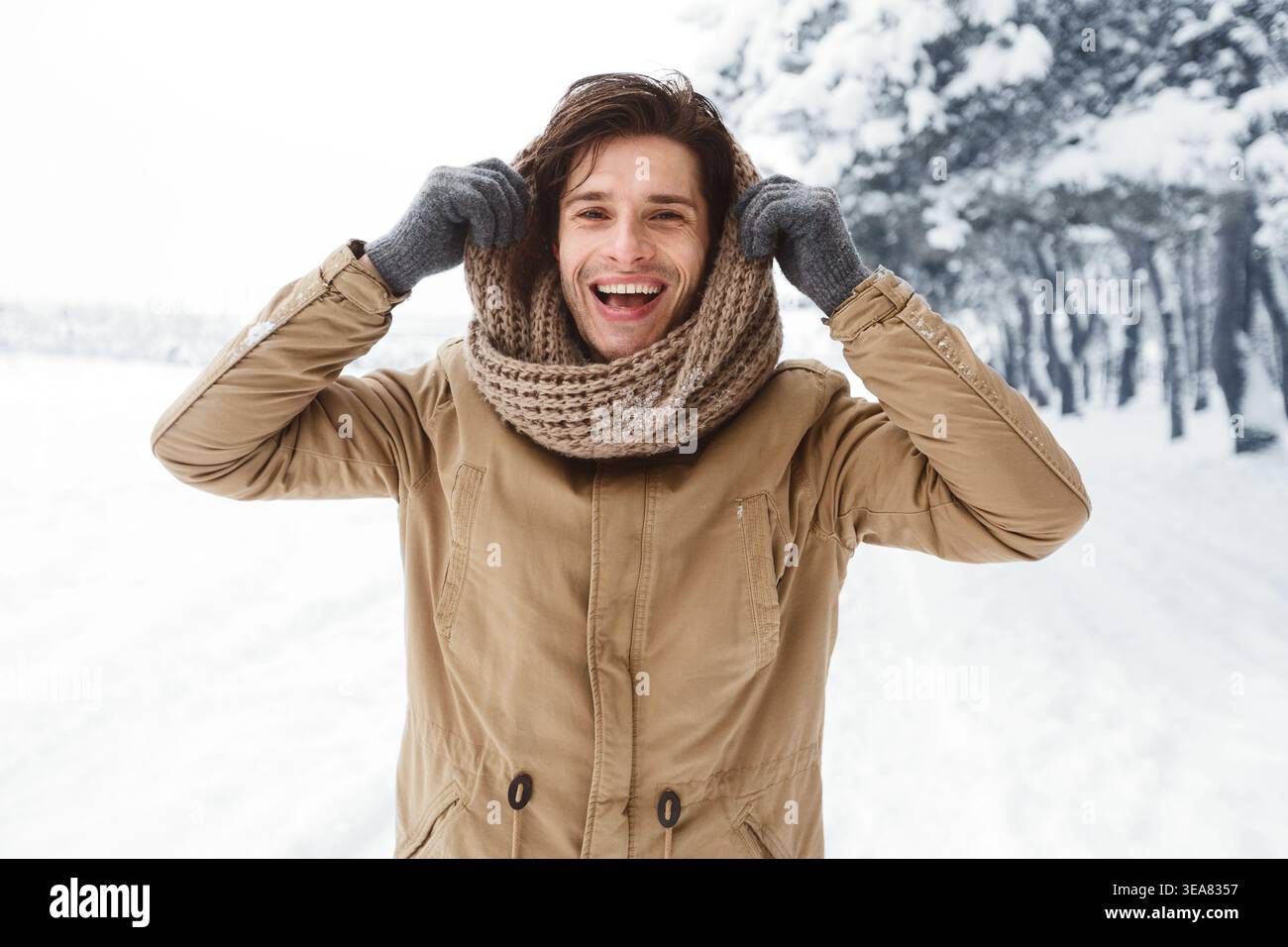 Handsome Man Pulling Scarf On Head Standing In Snowy Forest Stock Photo