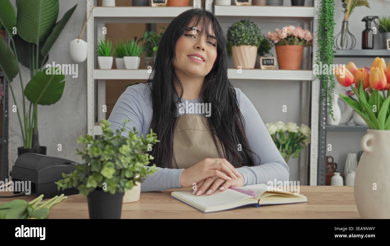 Woman florist in apron smiling with hands clasped on open notebook at studio counter, surrounded by potted plants, vases, shelves and tulips; welcomin Stock Photo