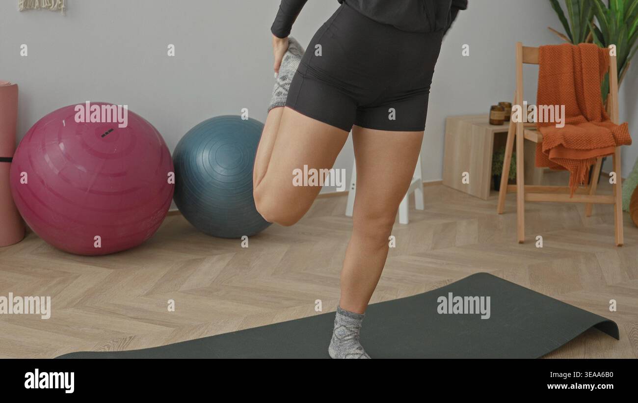 Young woman balancing on one leg on a yoga mat in a studio with visible leg skin, exercise balls, chair and plant nearby; balance focus. Stock Photo