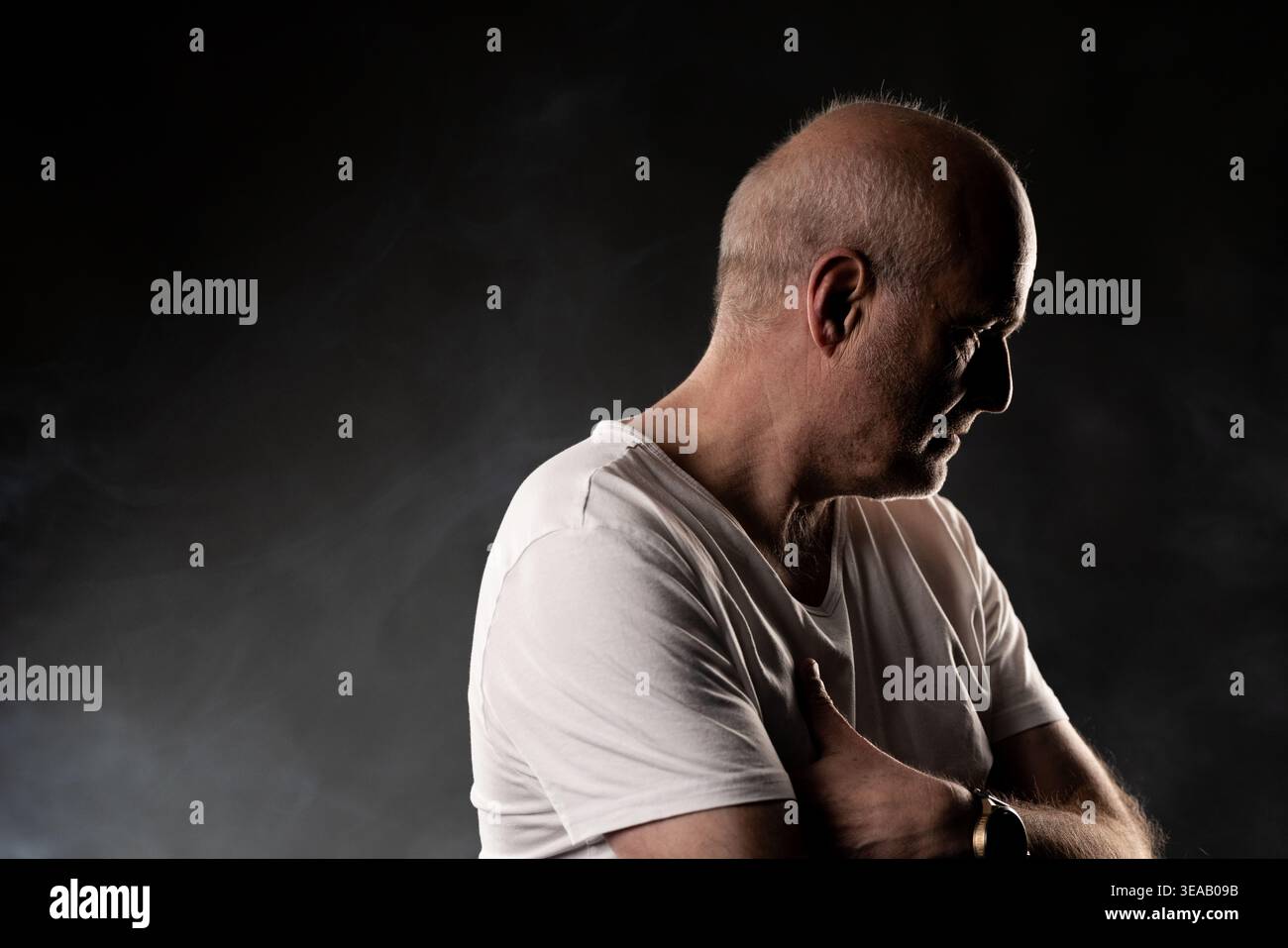 Mature bald man in profile looking down, contemplating in dark setting Stock Photo