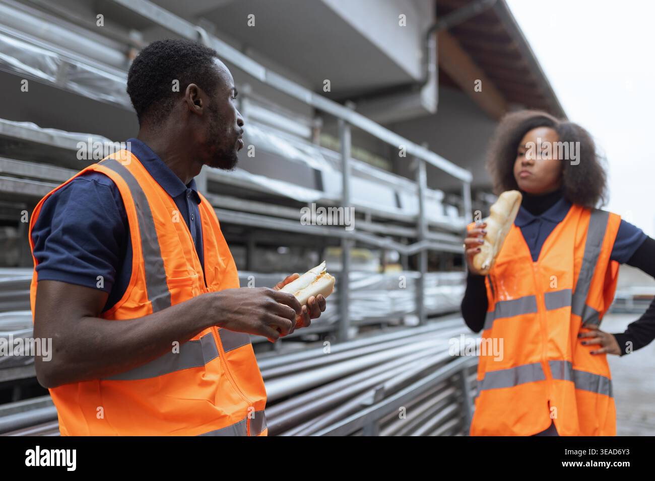 Two coworkers wearing reflective vests having fun during a lunch break, eating sandwiches outdoors at an industrial site. Stock Photo