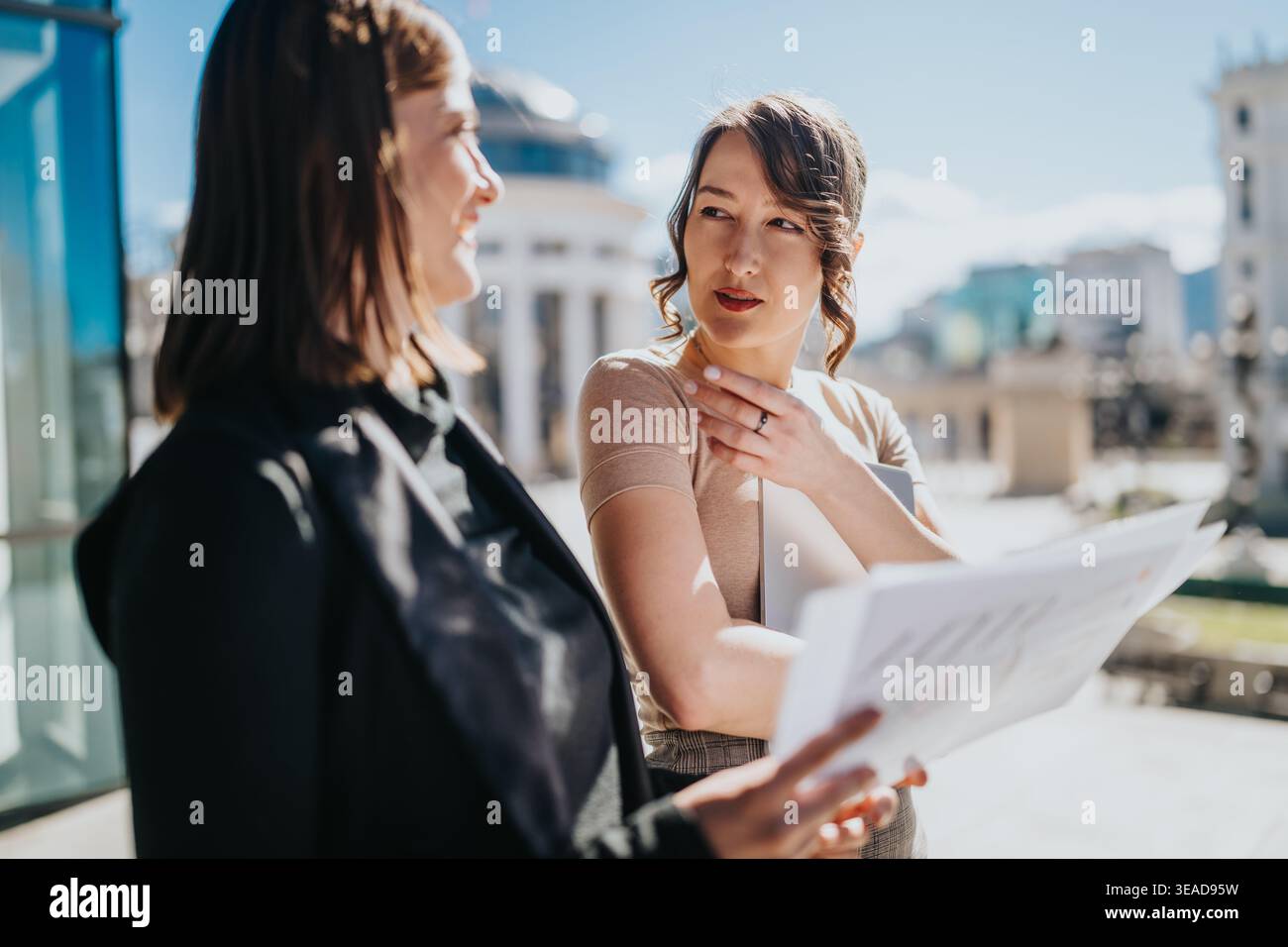 Two businesswomen discussing documents outside an office building during a daytime city meeting Stock Photo