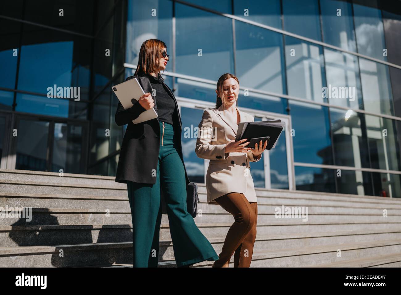 Two businesswomen walking down steps outside a modern office building discussing documents Stock Photo