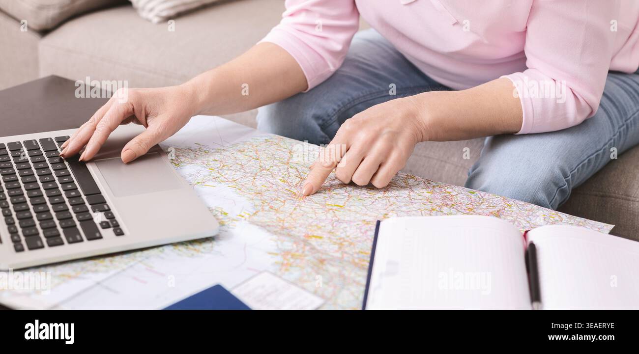 Person Using Laptop While Looking at Map in Living Room With Notebook Open on Table Stock Photo