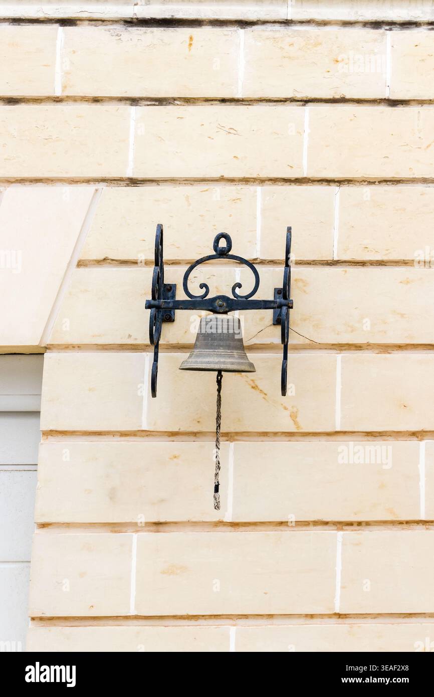 Vintage Iron Bell Mounted On Beige Stone Wall With Rope Pull And Rustic Bracket Stock Photo