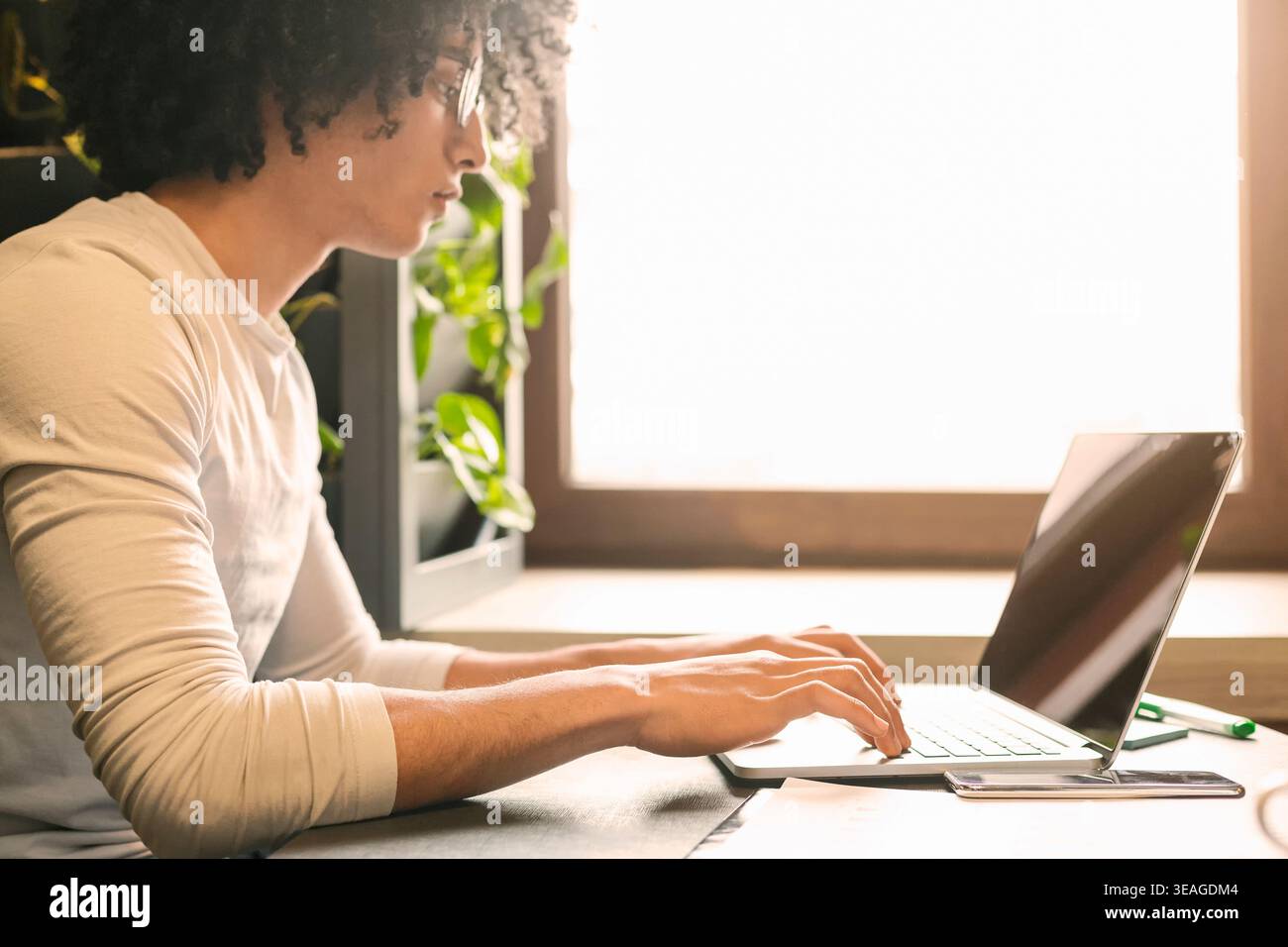 Young Man Working on Laptop by Window With Sunlight in a Cozy Indoor Space Stock Photo