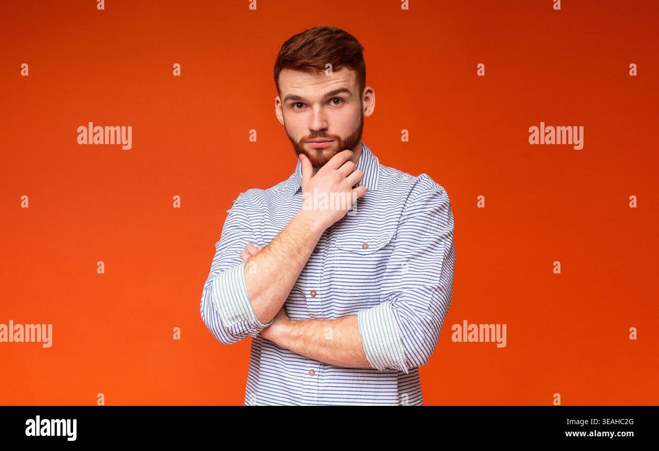 Man in Striped Shirt Poses Against Orange Background With Thoughtful Expression Stock Photo