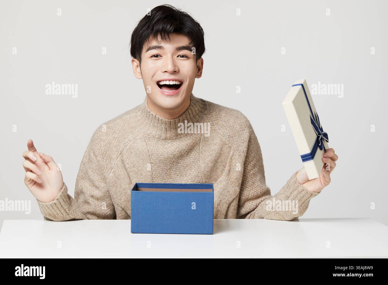 A young Asian man excitedly opens a gift box on a white table. Stock Photo