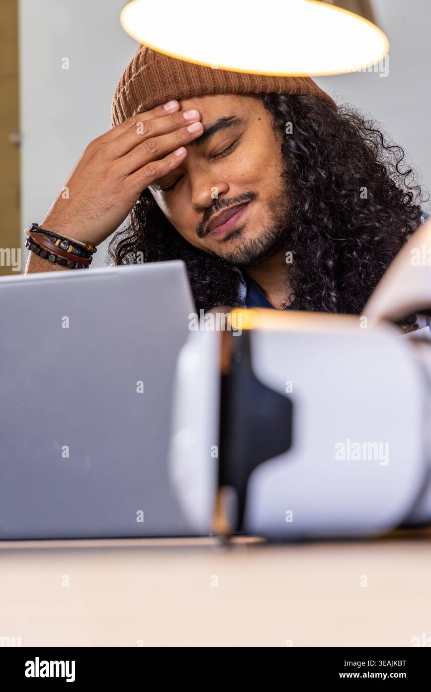 Man rubbing forehead, sitting at desk in home office wearing beanie with laptop and VR headset Stock Photo