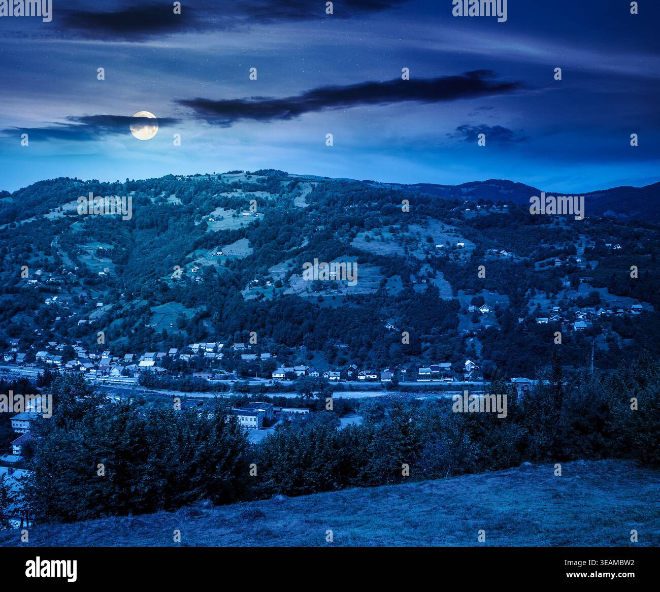 carpathian mountain landscape on summer night. full moon over view in to the valley of town near forested hill of mountain range. mysterious travel ba Stock Photo