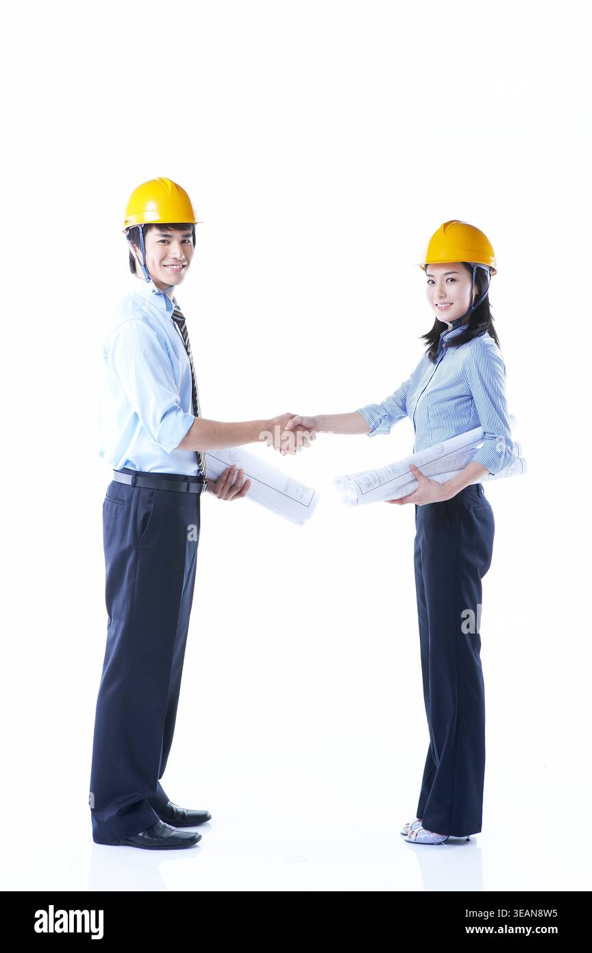 Two engineers in hard hats shake hands while holding blueprints, standing against a white background. Stock Photo