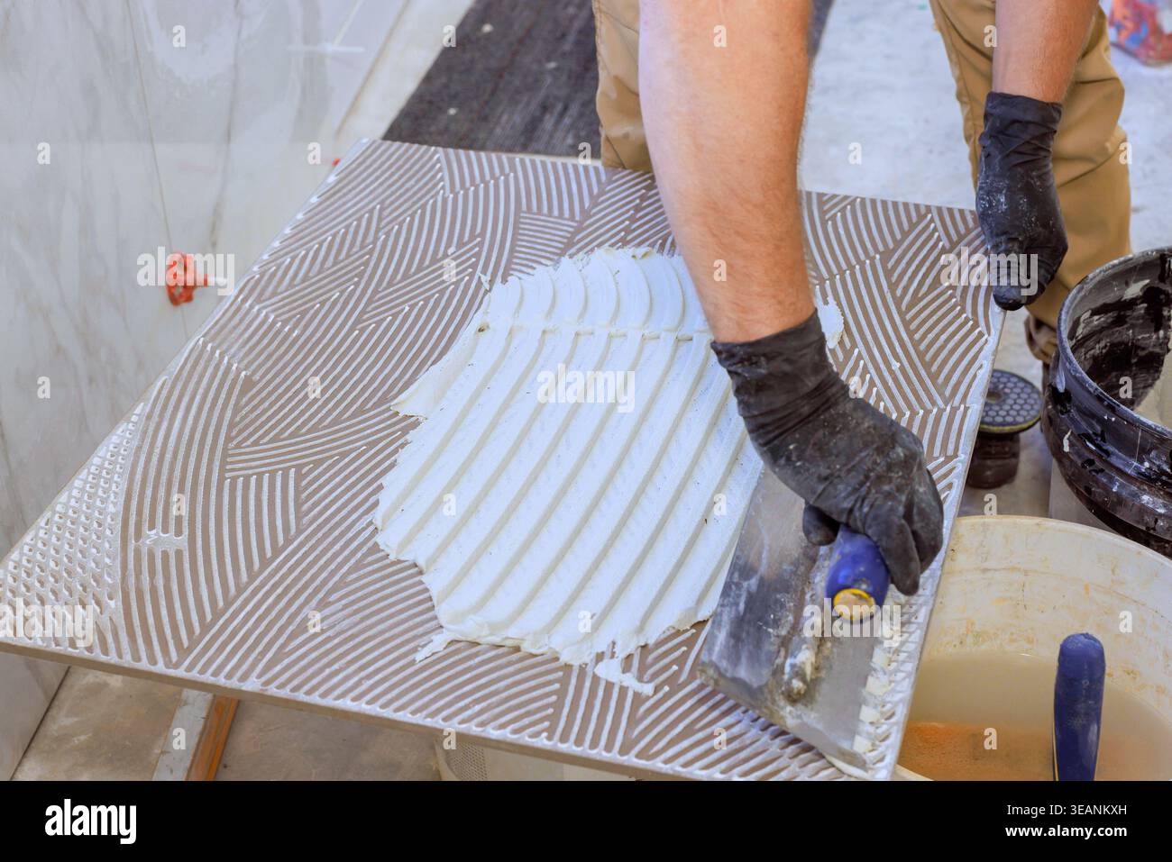 Tiling worker uses trowel to spread adhesive on tile during flooring project in construction area. Stock Photo