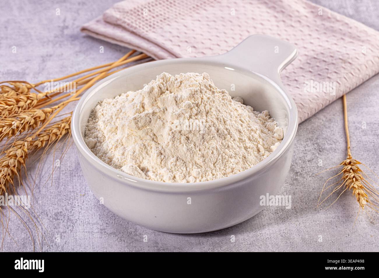 Close up still life image featuring ceramic bowl filled with fresh white flour, golden wheat stalks, folded light pink kitchen towel Stock Photo
