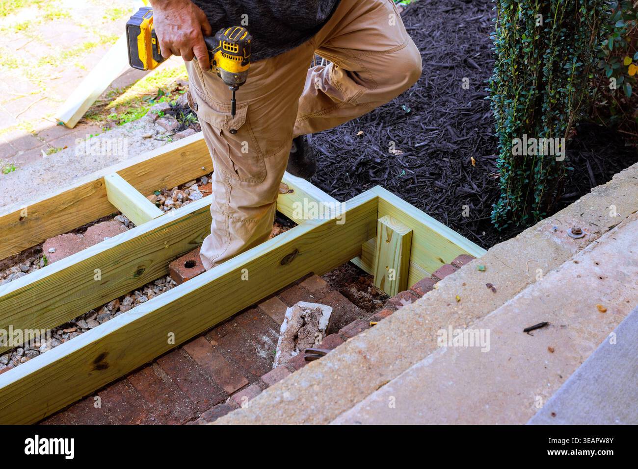 Worker is using drill to secure steps wood pieces while standing on step outside, surrounded by construction materials. Stock Photo
