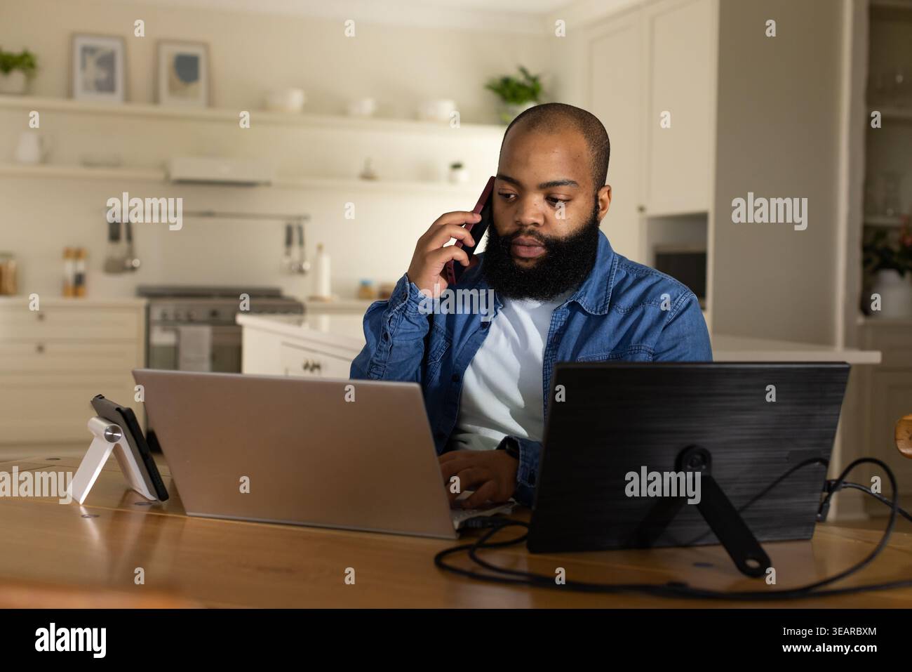 Bearded man working in kitchen using dual screens and speaking on phone stand, in denim jacket. Remote, laptops, cables, plants, modern, homeoffice, p Stock Photo