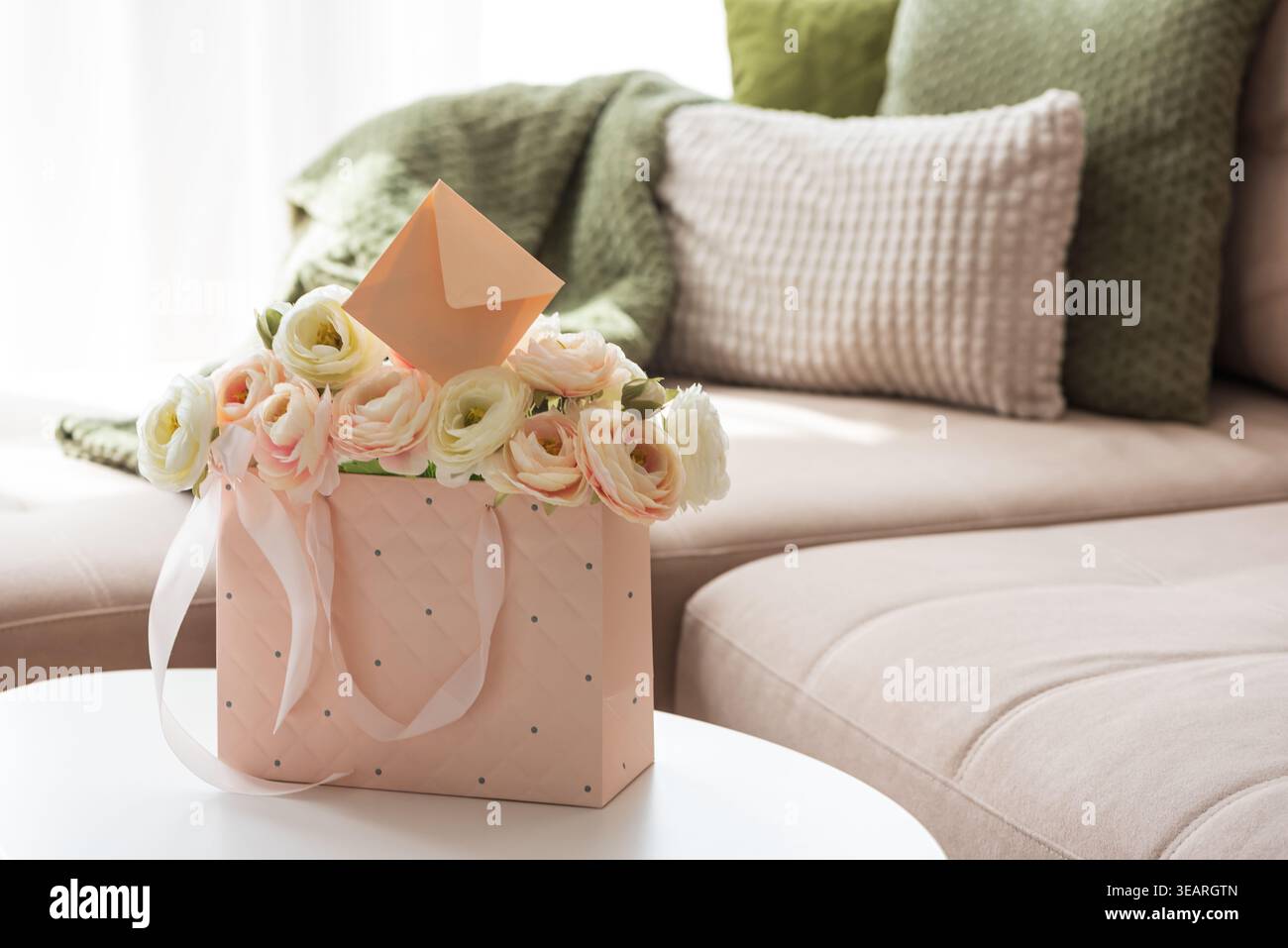 Delicate lifestyle composition featuring a quilted pink gift bag filled with cream and blush ranunculus flowers, accented by ribbon handles and decora Stock Photo